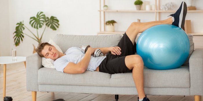 laziness and workout tired guy sleeping on couch with dumbbell and fitness ball, in living room interior