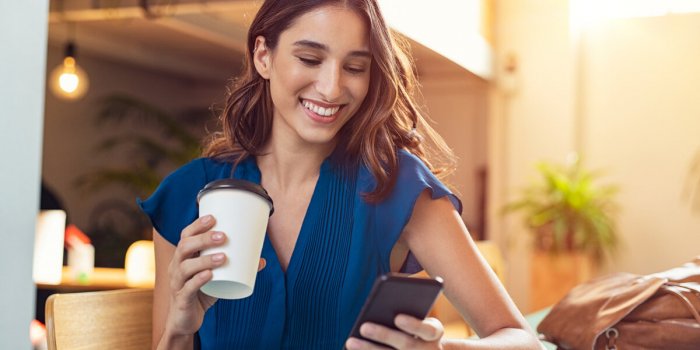 young beautiful woman holding coffee paper cup and looking at smartphone while sitting at cafeteria happy university stud...