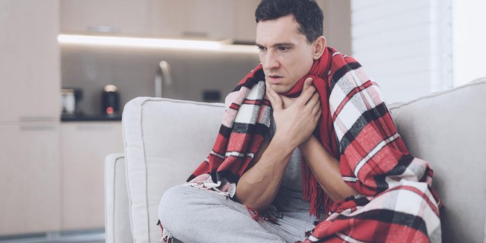 a man with a cold sits on the couch, hiding behind a red rug he's holding his throat because it hurts badly