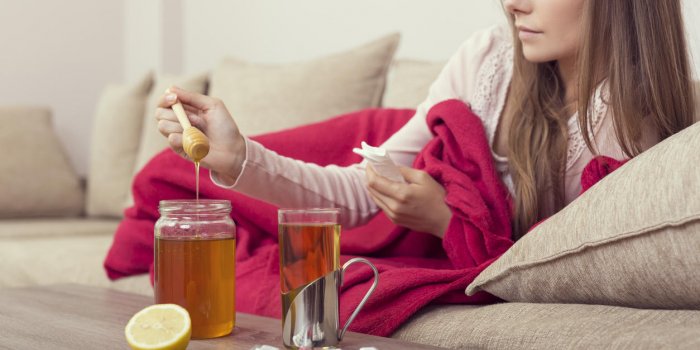 sick woman lying in bed with high fever and a flu, putting some honey in her tea lemon, honey jar and pills set around th...