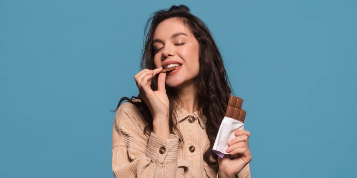 glad happy millennial european woman with closed eyes enjoys taste of chocolate bar, isolated on blue background, studio ...